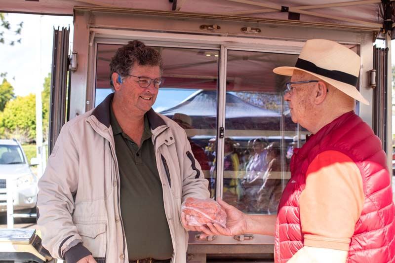 Two men in a tent booth exchange an item and chat outdoors at a market stall