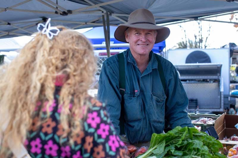 Smiling vendor in cowboy hat behind a market stall, serving greens to a customer with curly blonde hair.