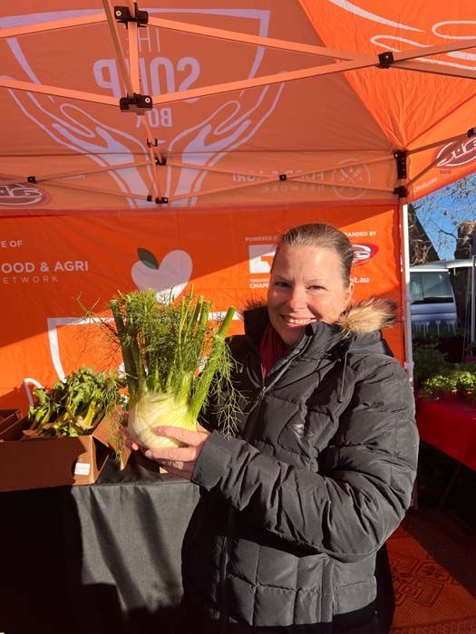 Woman holding a bundle of fresh greens at an orange market stall, smiling in bright sunlight