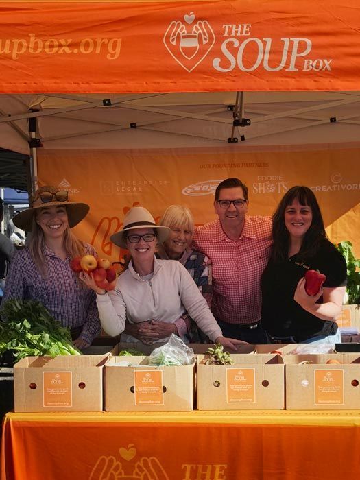 People at an orange FOOD & AGRI market tent displaying potted plants outdoors