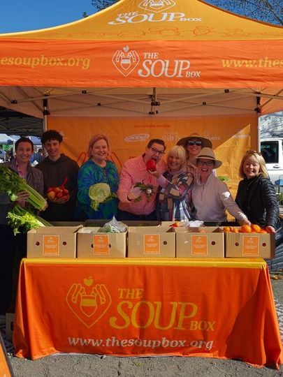 People at an orange FOOD & AGRI market tent displaying potted plants outdoors