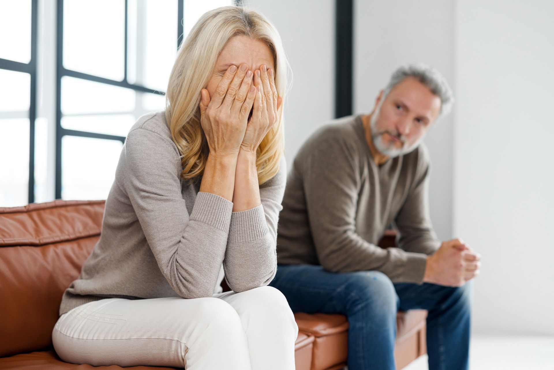 Woman covering face, sitting on couch, man sits nearby, both appear distressed.
