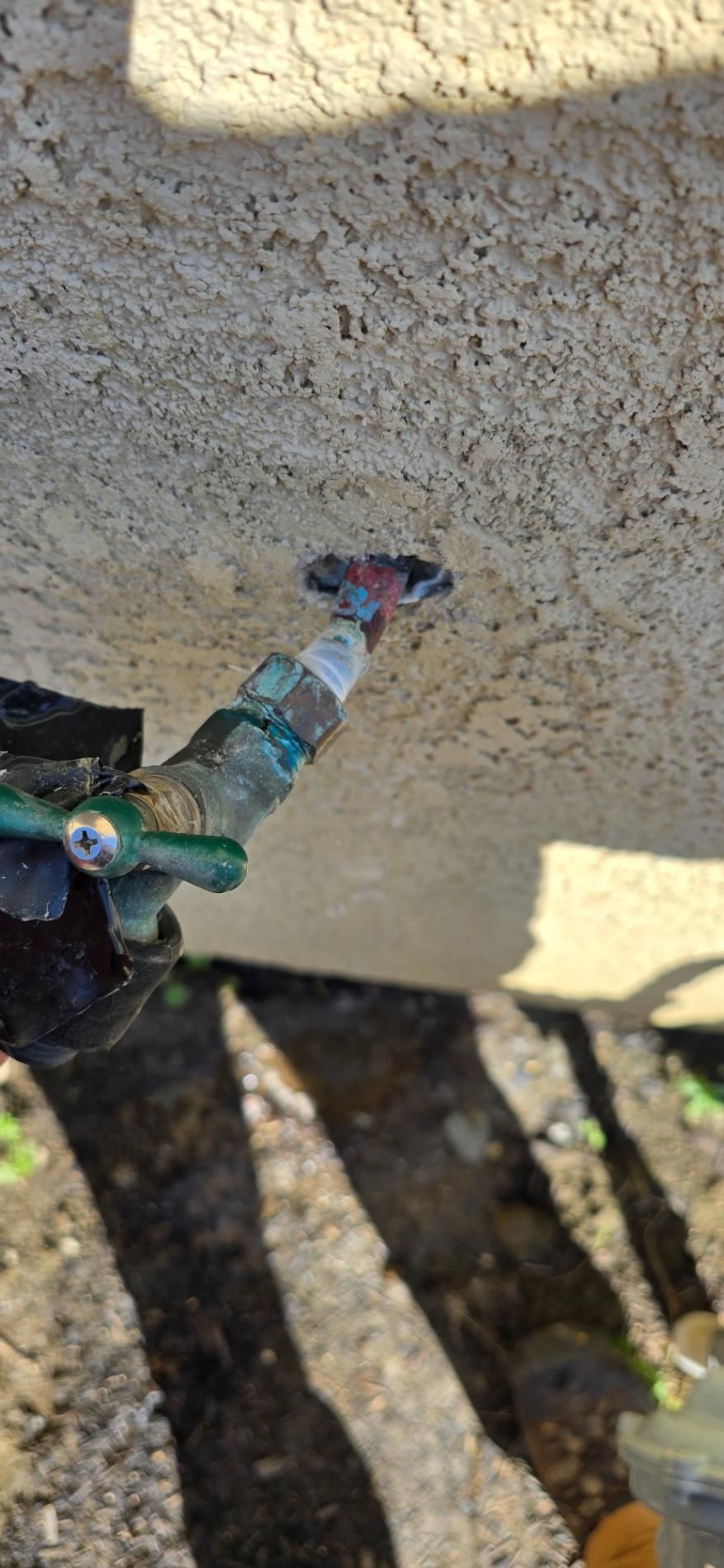 A garden hose sprayer aimed at a stucco wall, with sunlight and shadows on the ground.
