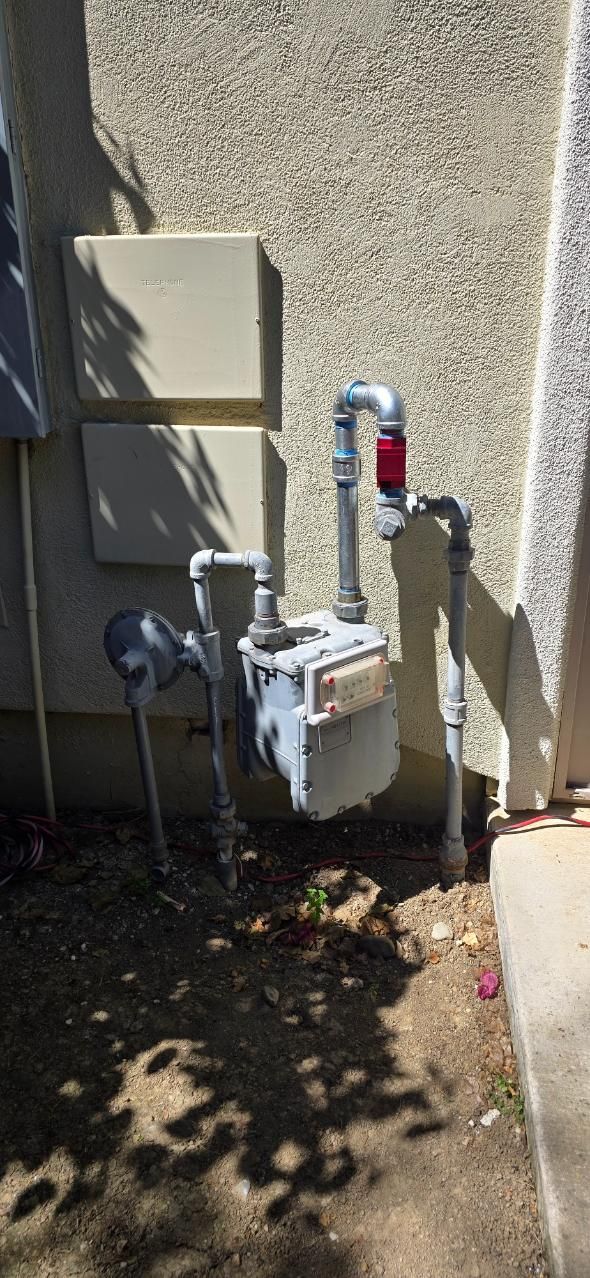 Outdoor utility meters and pipes mounted on a stucco wall beside a small electrical box.