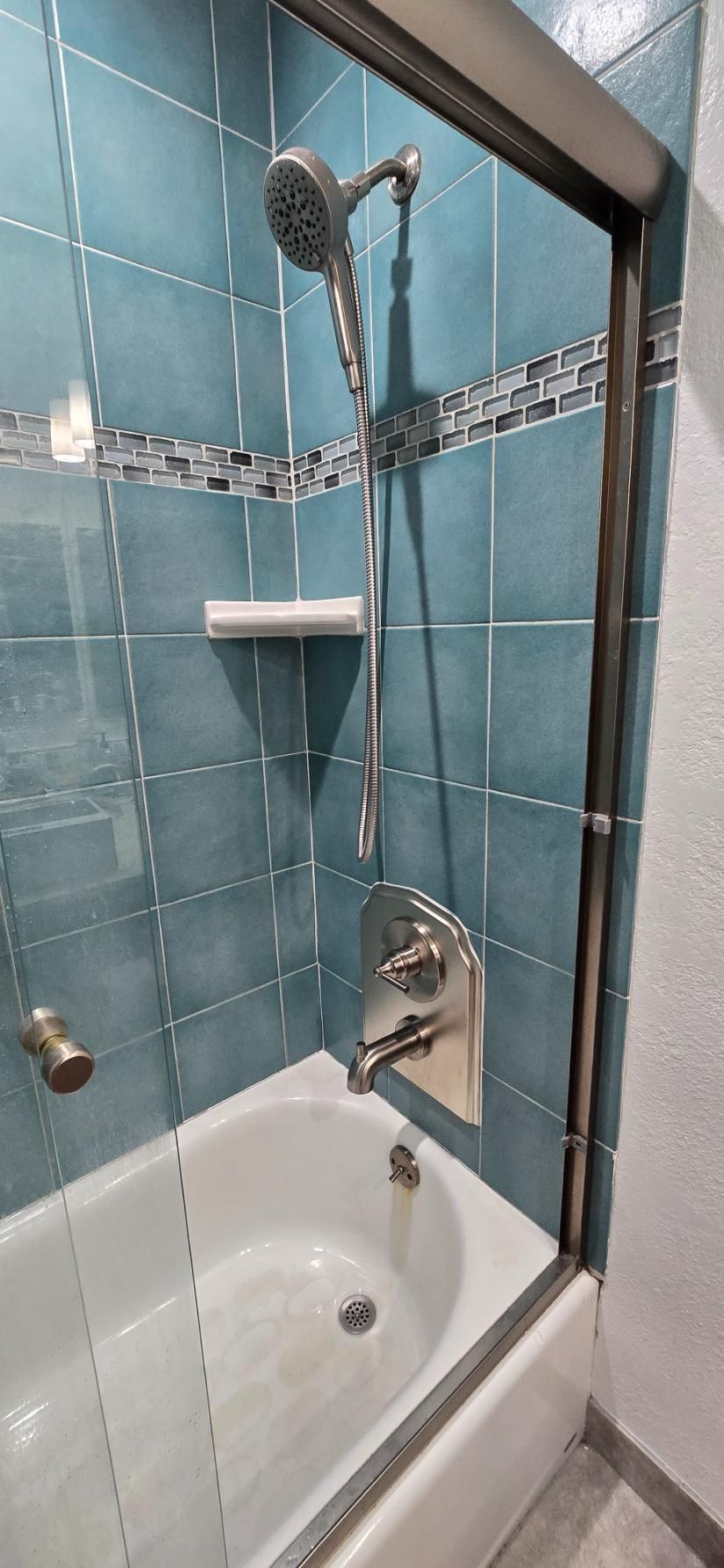 Small shower stall with teal tile, glass door, showerhead, hose, and bathtub edge