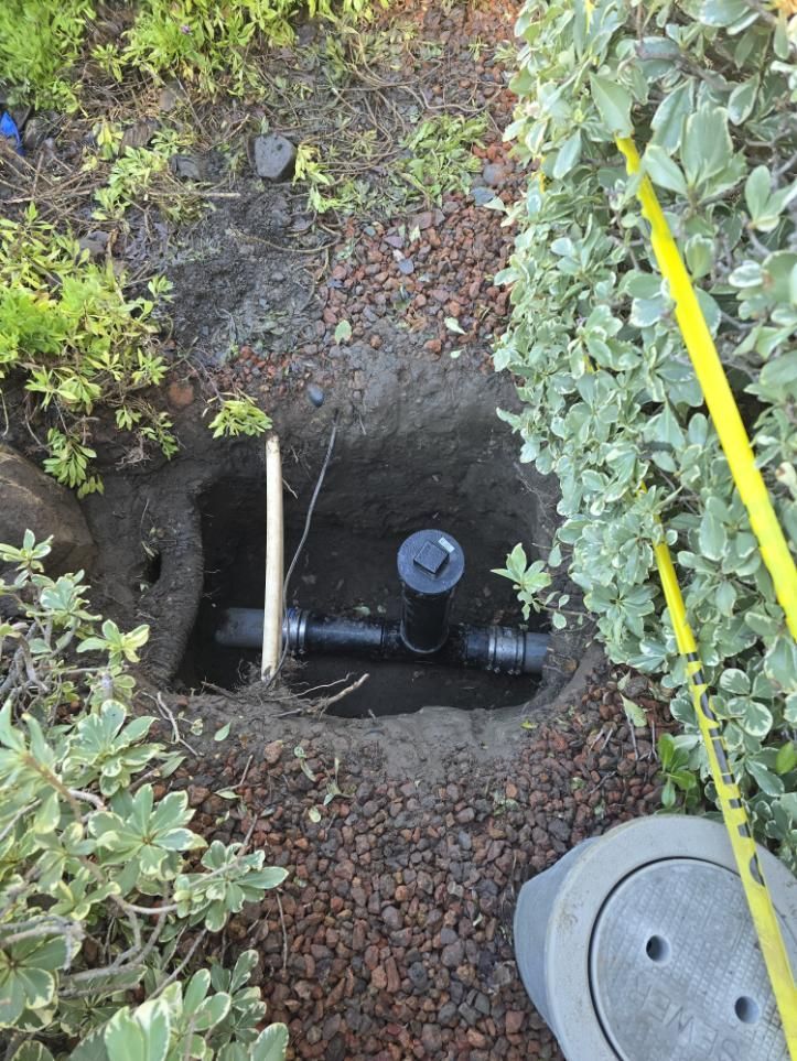 Open hole in garden with black pipe, valve, and yellow marker near shrubs and gravel