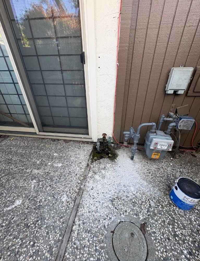 Outdoor utility area beside a house wall with gas meters, pipes, gravel, and a bucket