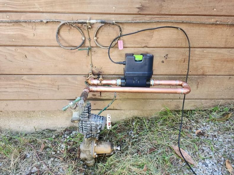 Water pump and pipes mounted on a wooden wall outdoors with cables and grass below