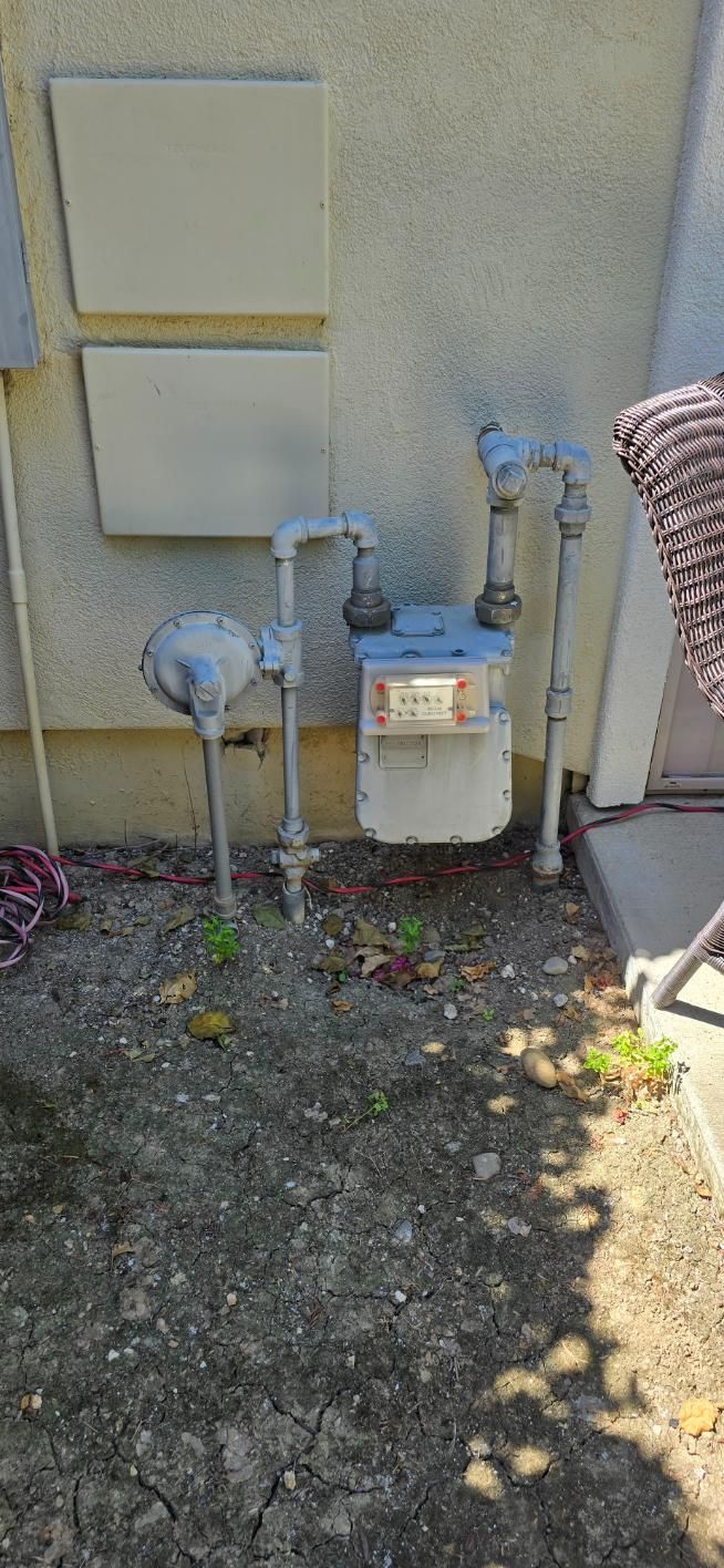 Outdoor utility meters and gas pipes mounted on a beige wall beside gravel ground