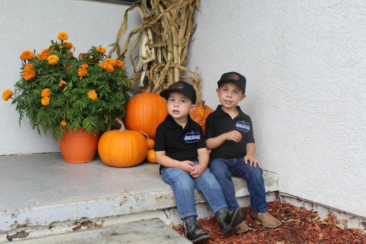Two children sitting on a porch beside pumpkins and orange flowers, with fall decorations behind them
