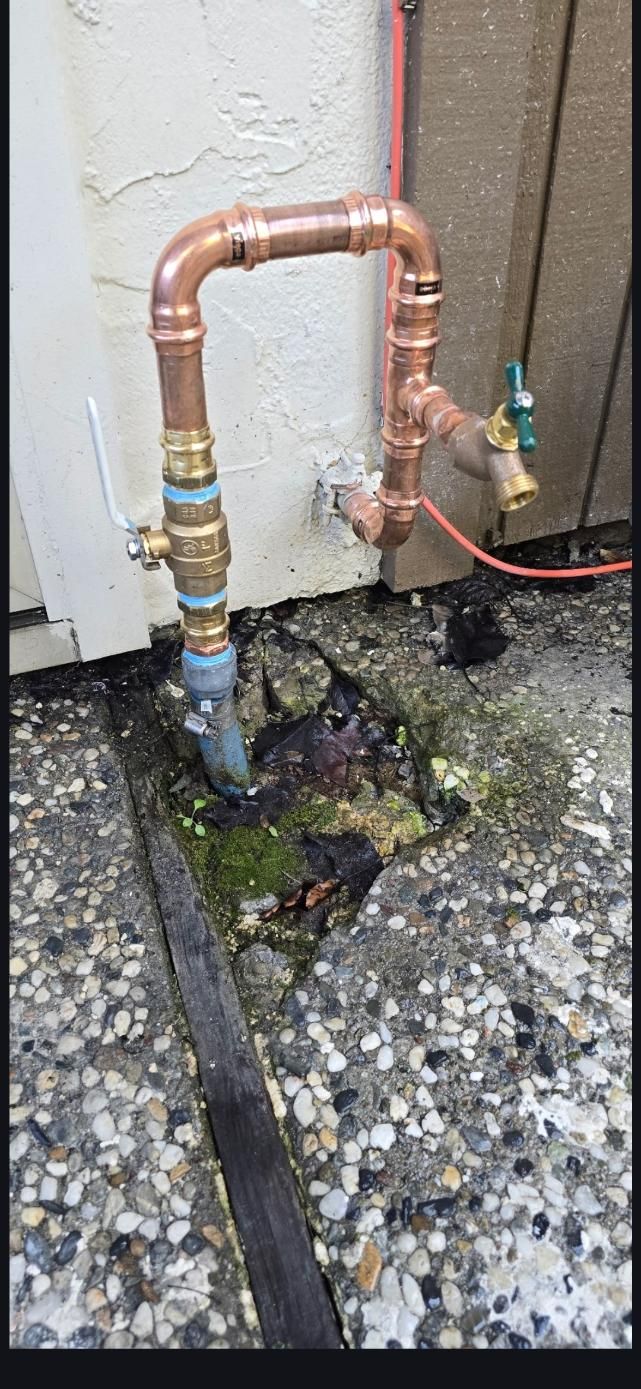 Copper pipes and a valve connected to an outdoor water line over a wet, cracked concrete surface.