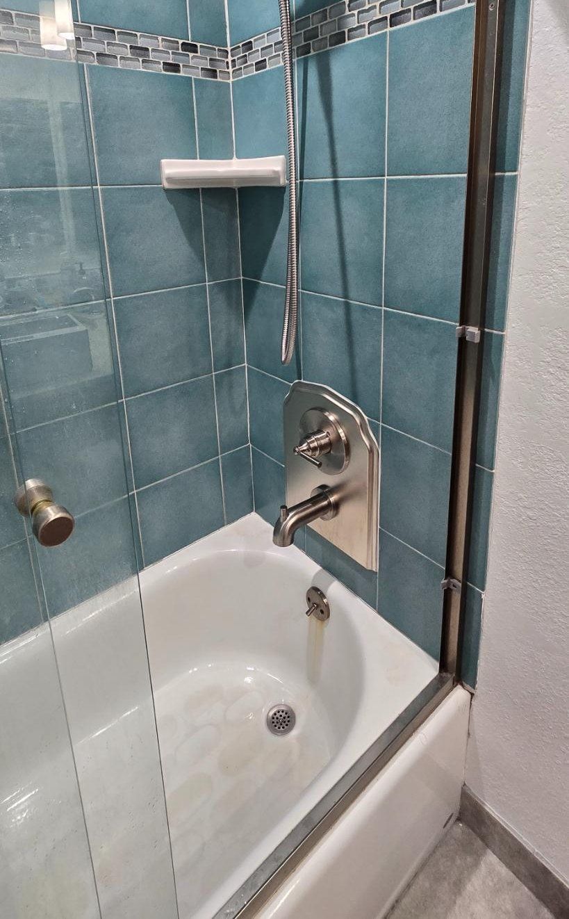 Small tiled shower with white tub, chrome faucet, handheld showerhead, and built-in soap shelf.