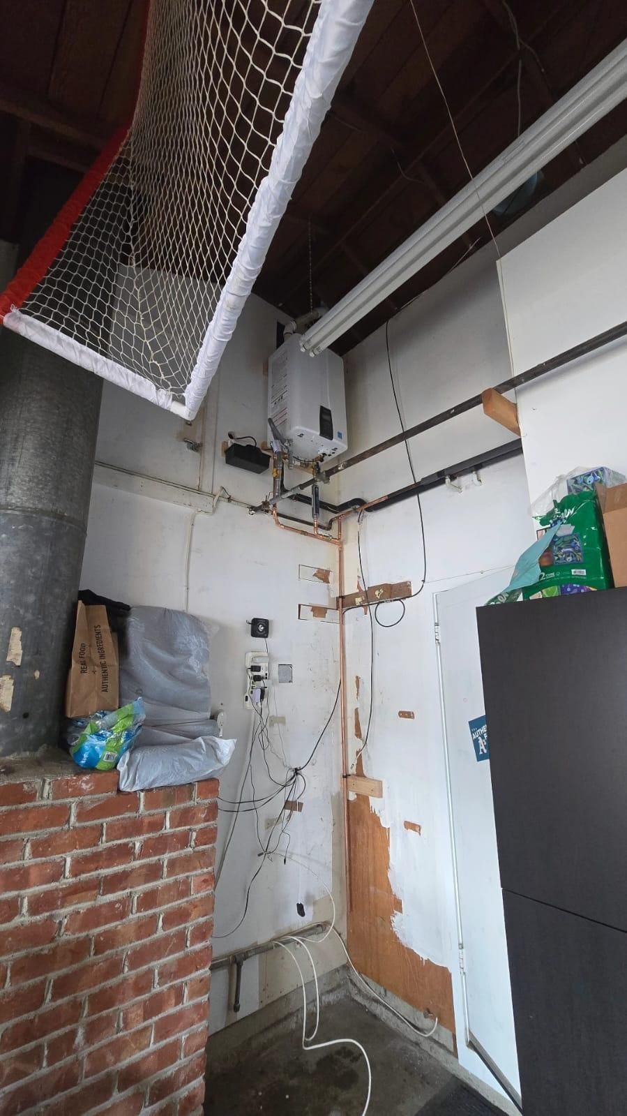 Utility room with white walls, brick partition, exposed pipes, and a door beneath a ceiling storage net.