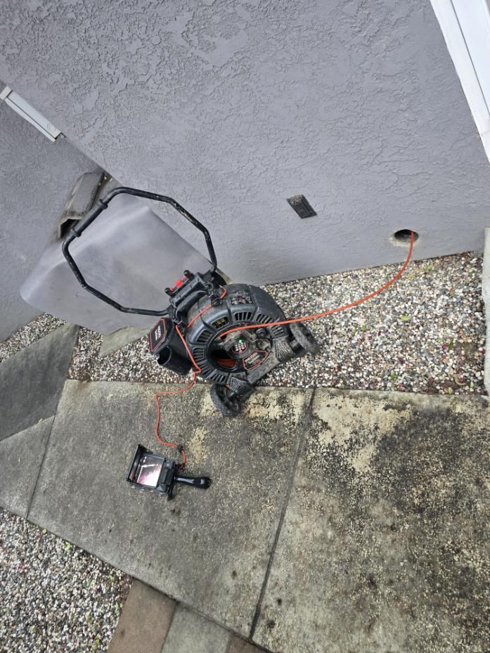 Damaged red-and-black drone on a sidewalk beside a wall and gravel strip, with a detached controller nearby
