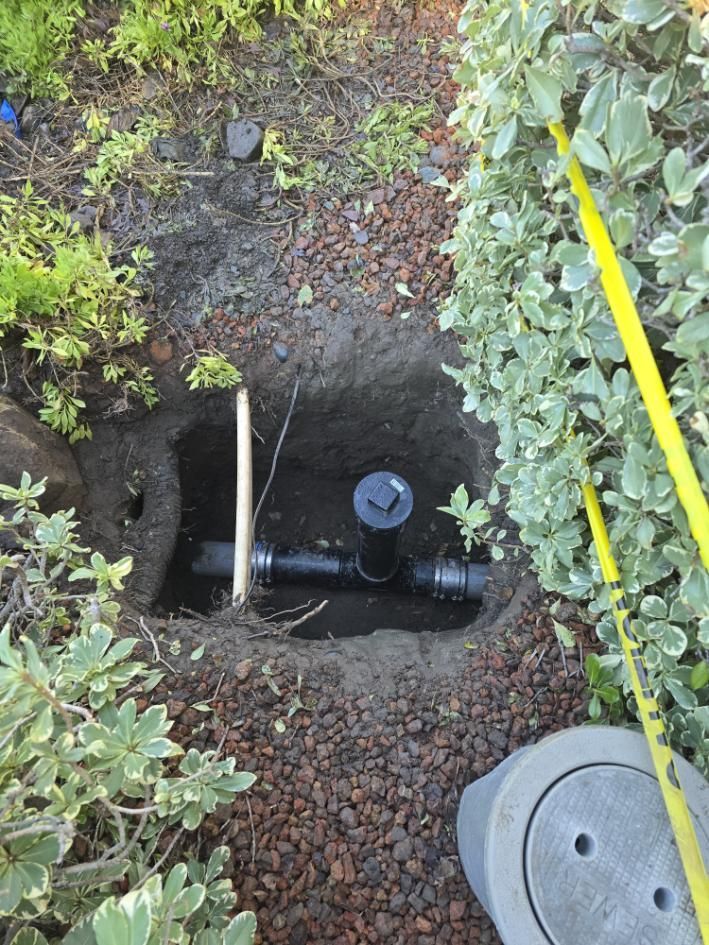 Open underground utility valve with black pipes in a gravel-lined hole, surrounded by greenery.