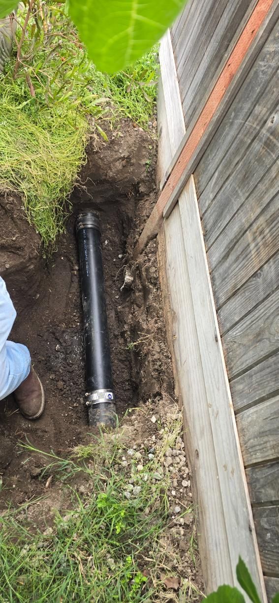 Open trench beside a fence and grass, exposing a black pipe with metal clamps.