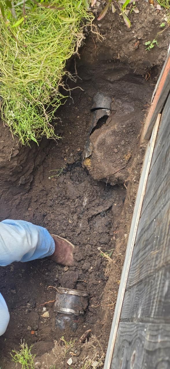 Worker inspecting muddy trench beside a wall, with exposed pipe and grass edge