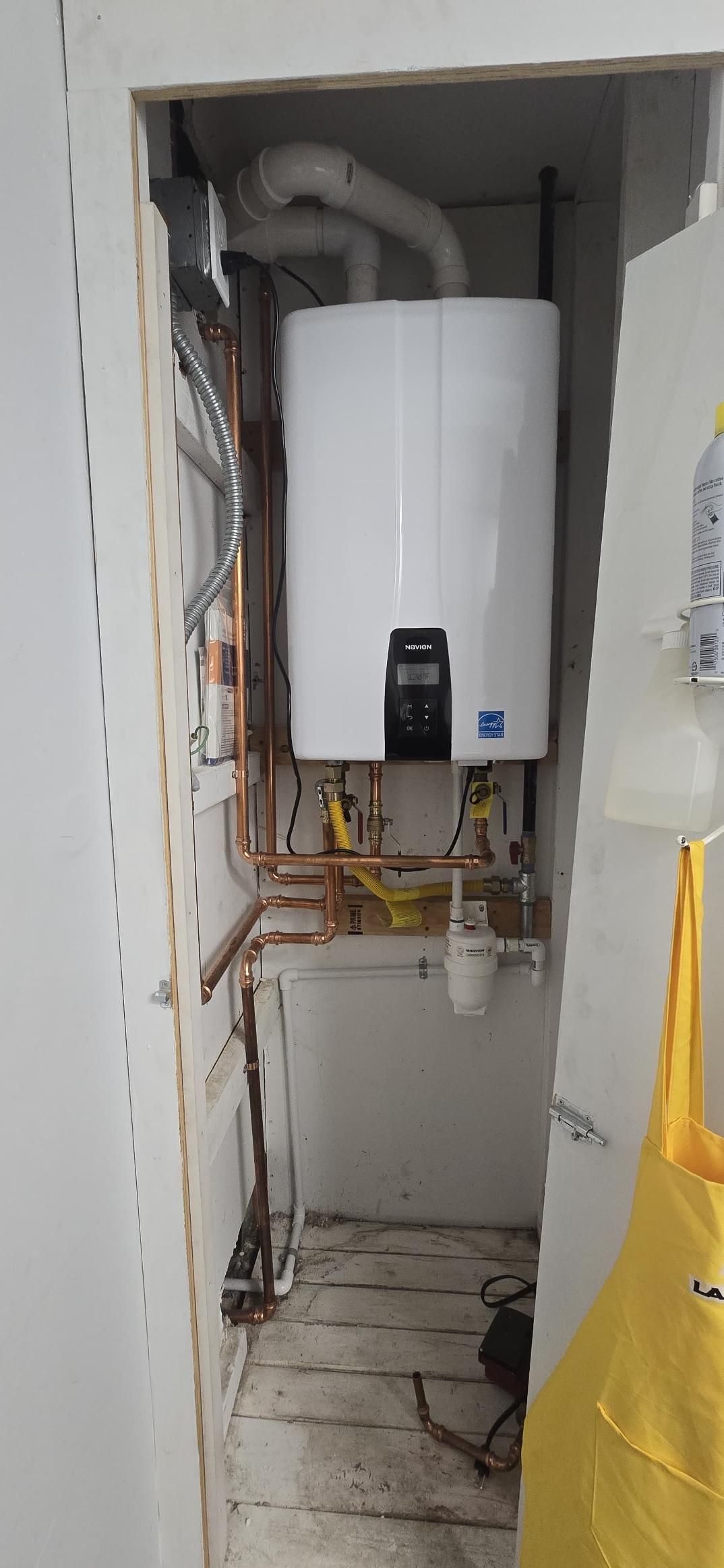 White water heater in a narrow utility closet with exposed pipes and wiring, and a yellow bag at right.
