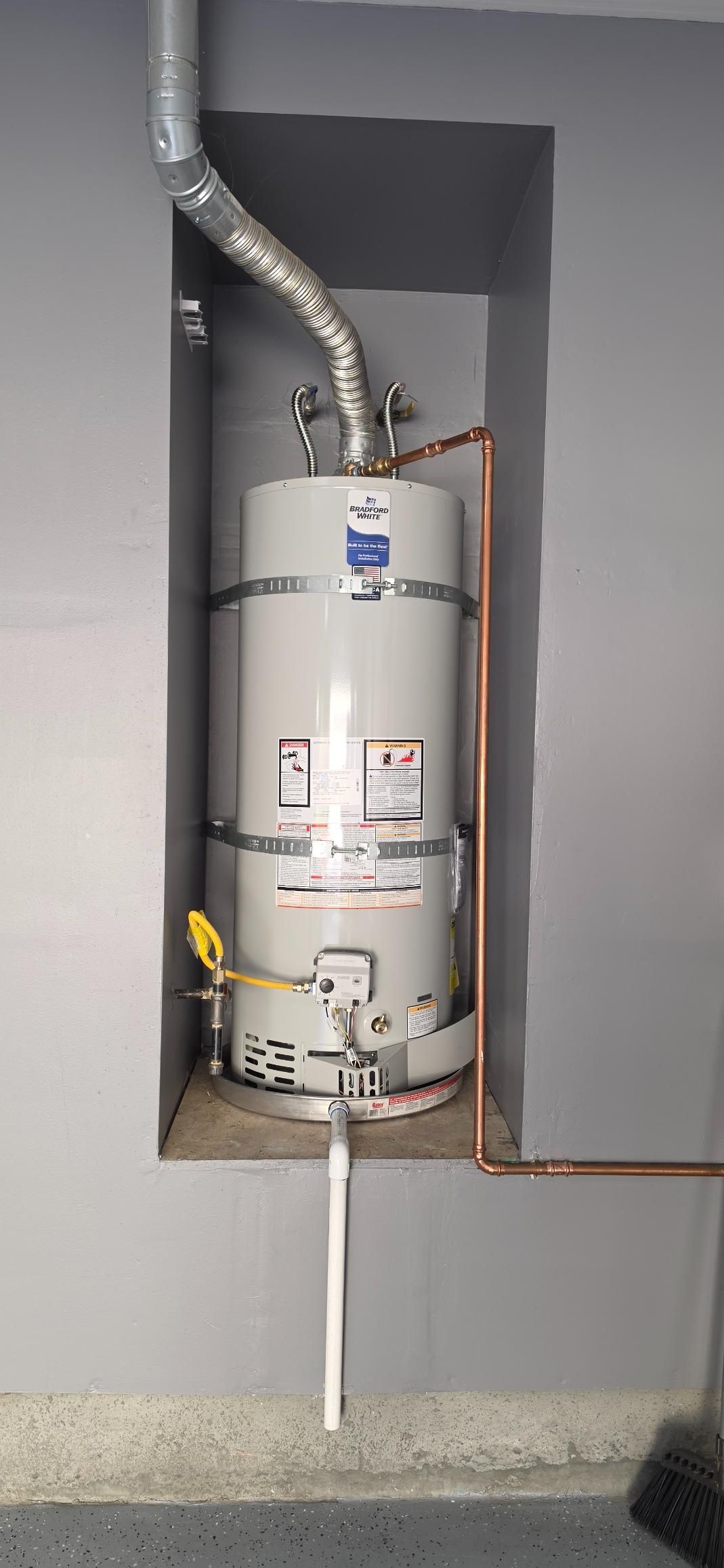 White water heater in a gray utility closet with exposed pipes and venting.