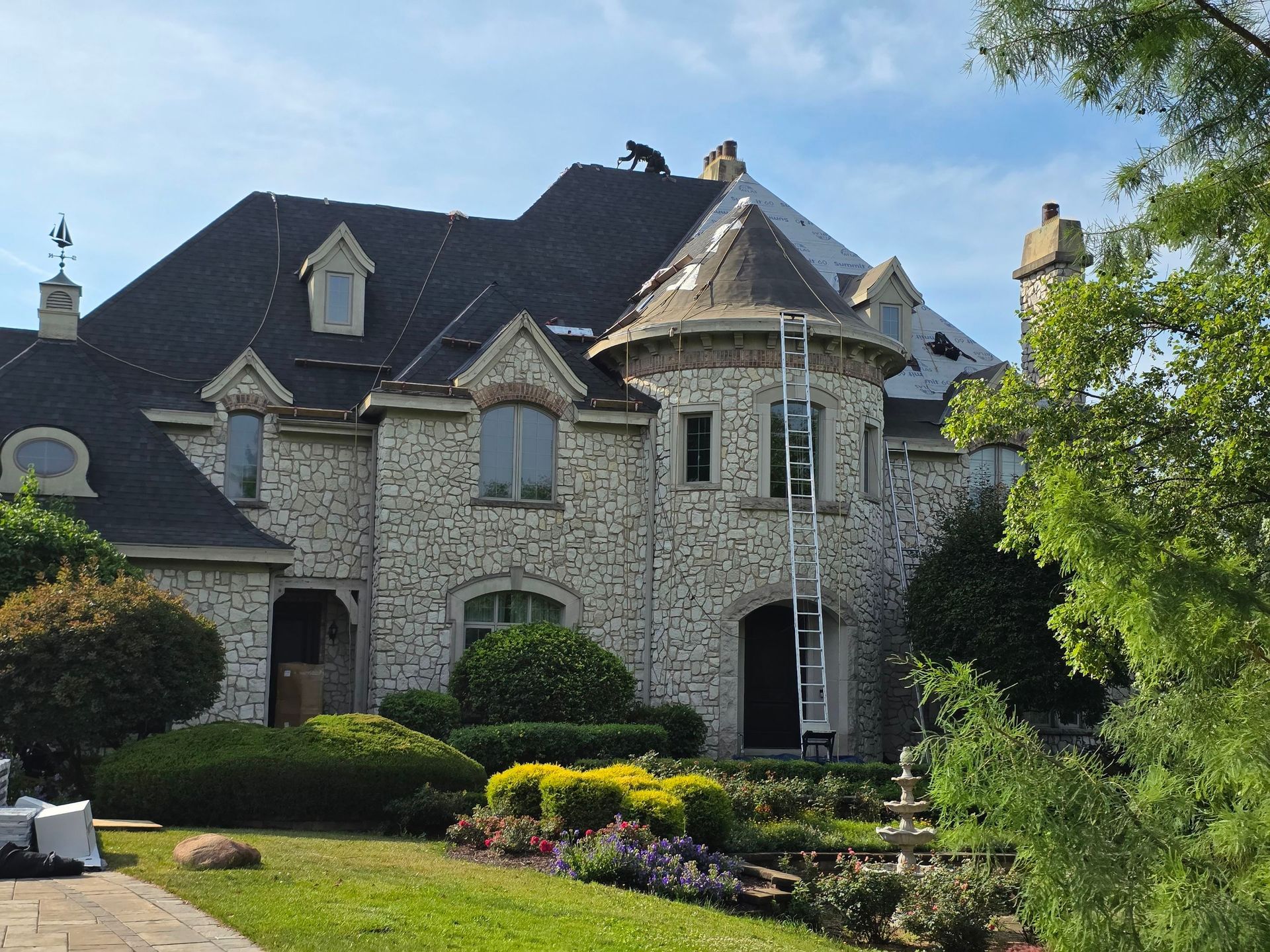 A stone-facade house with a turret and dark shingle roof, currently undergoing roof repairs with a ladder leaning against it.
