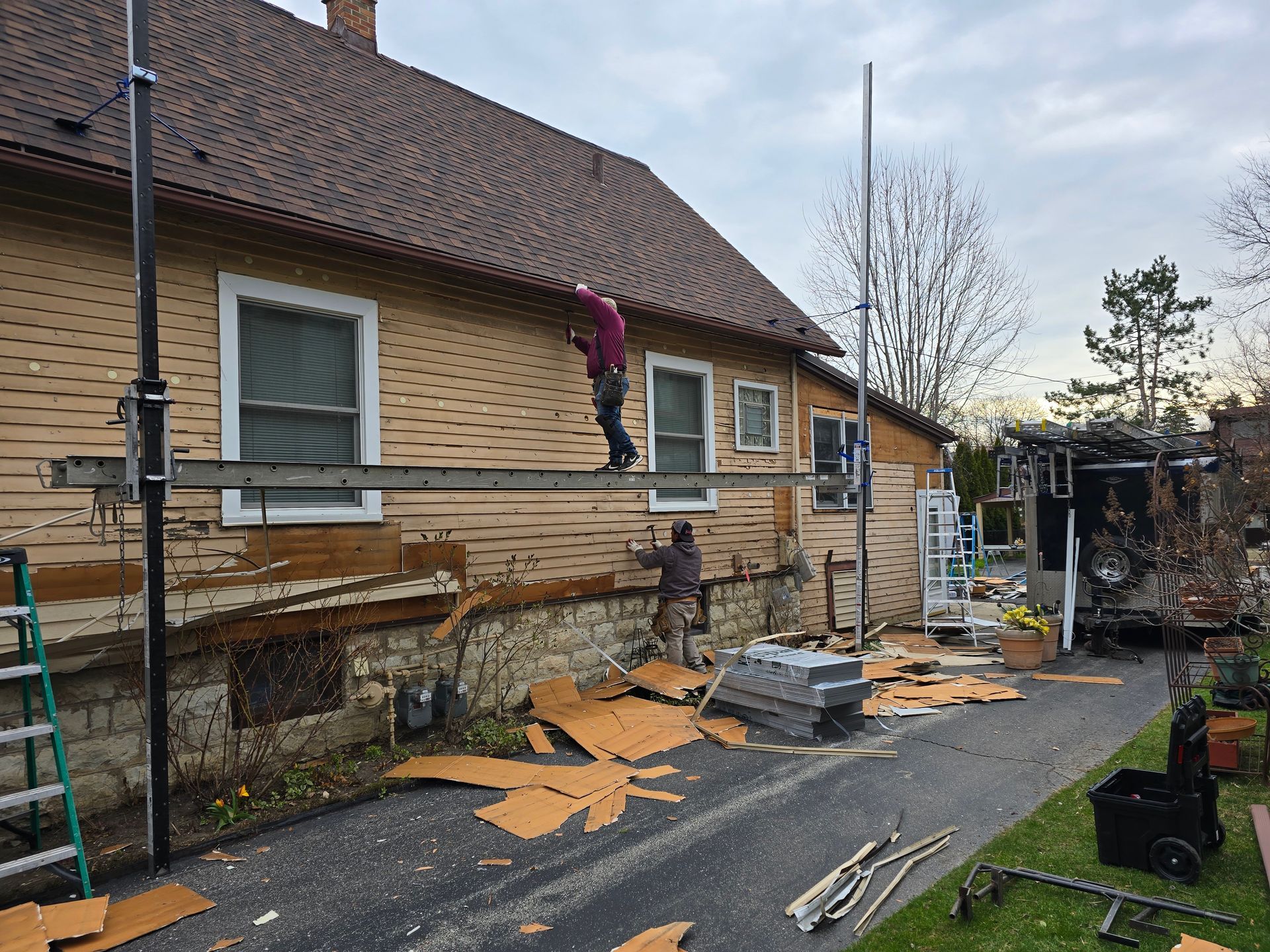Two workers remove exterior siding from a house while standing on a suspended scaffold.