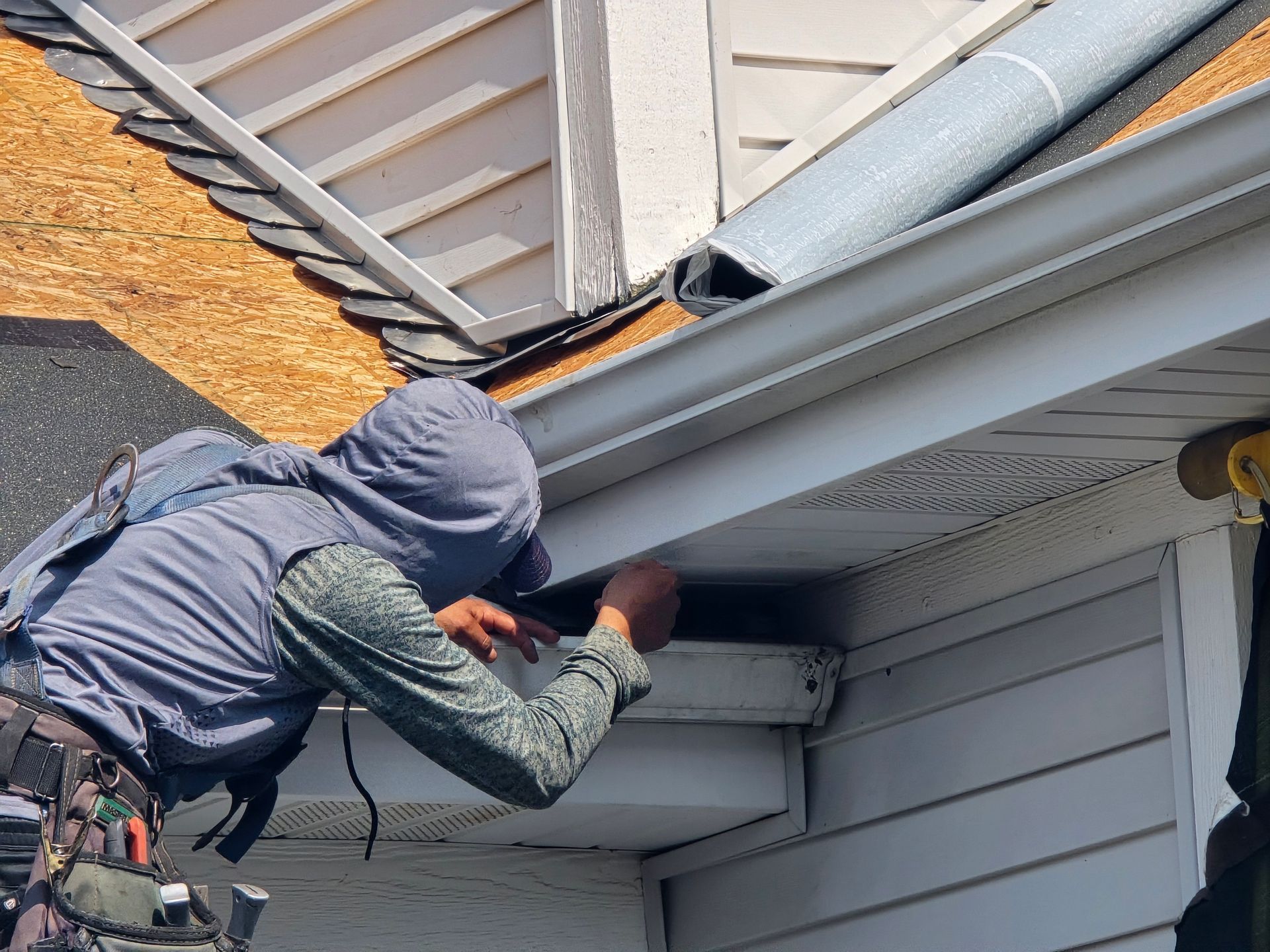 A person in a hooded gray vest and long-sleeved shirt works on the soffit and gutter of a residential roof.