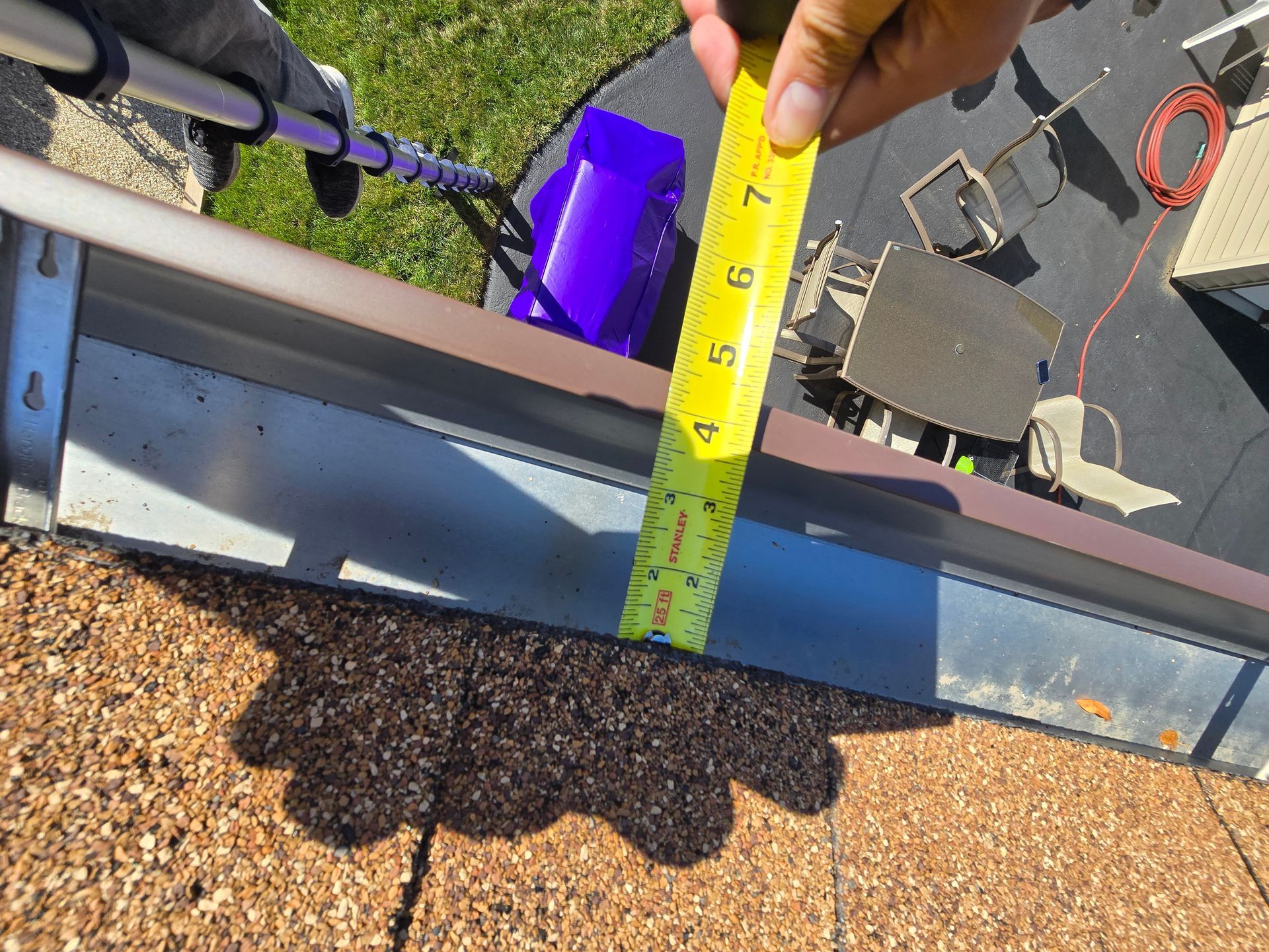 A hand holds a yellow tape measure against a roof gutter to show its depth, with a driveway and patio furniture below.