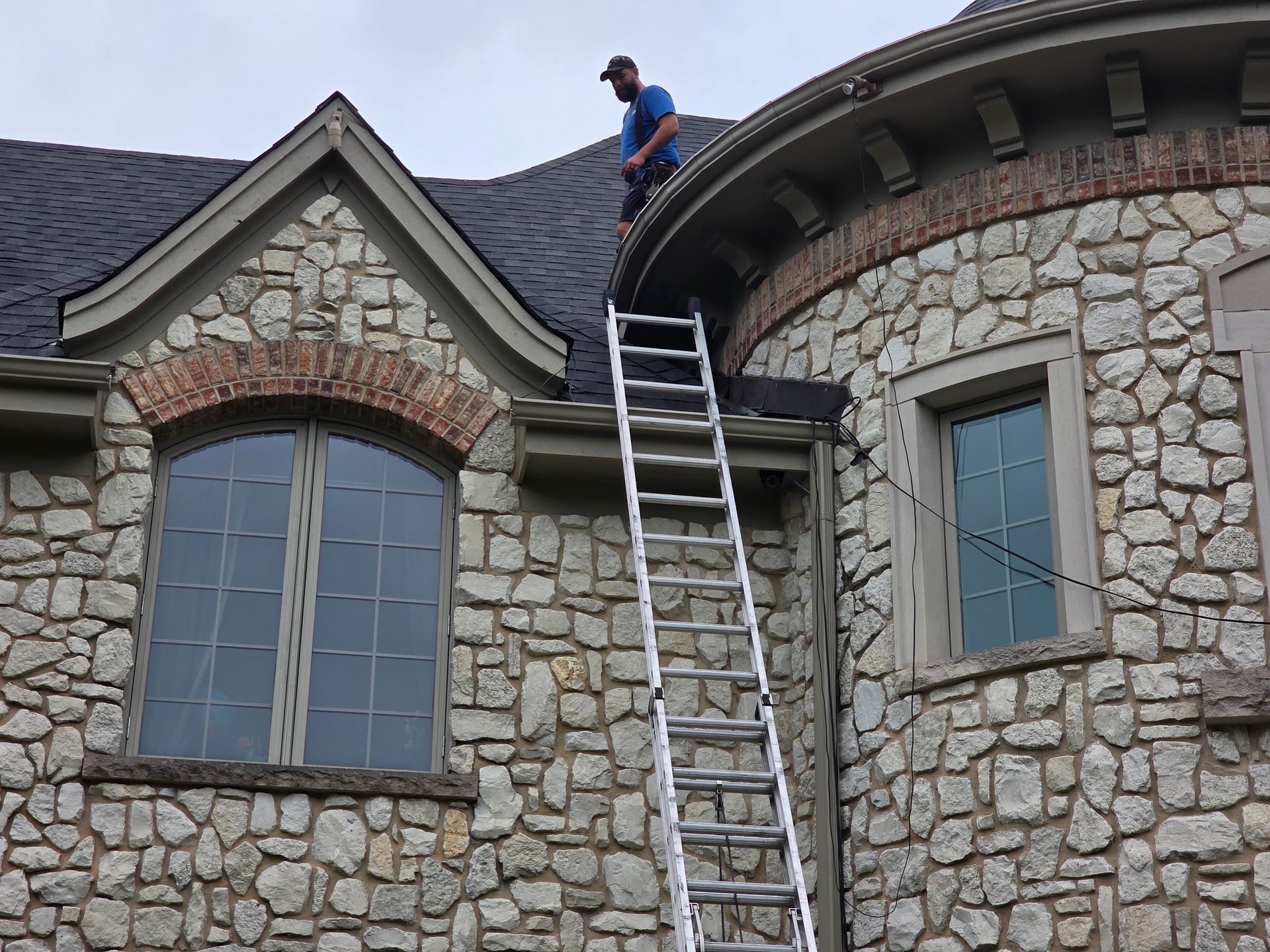 A person stands on a roof near a tall ladder propped against a stone house with arched windows and a turret.