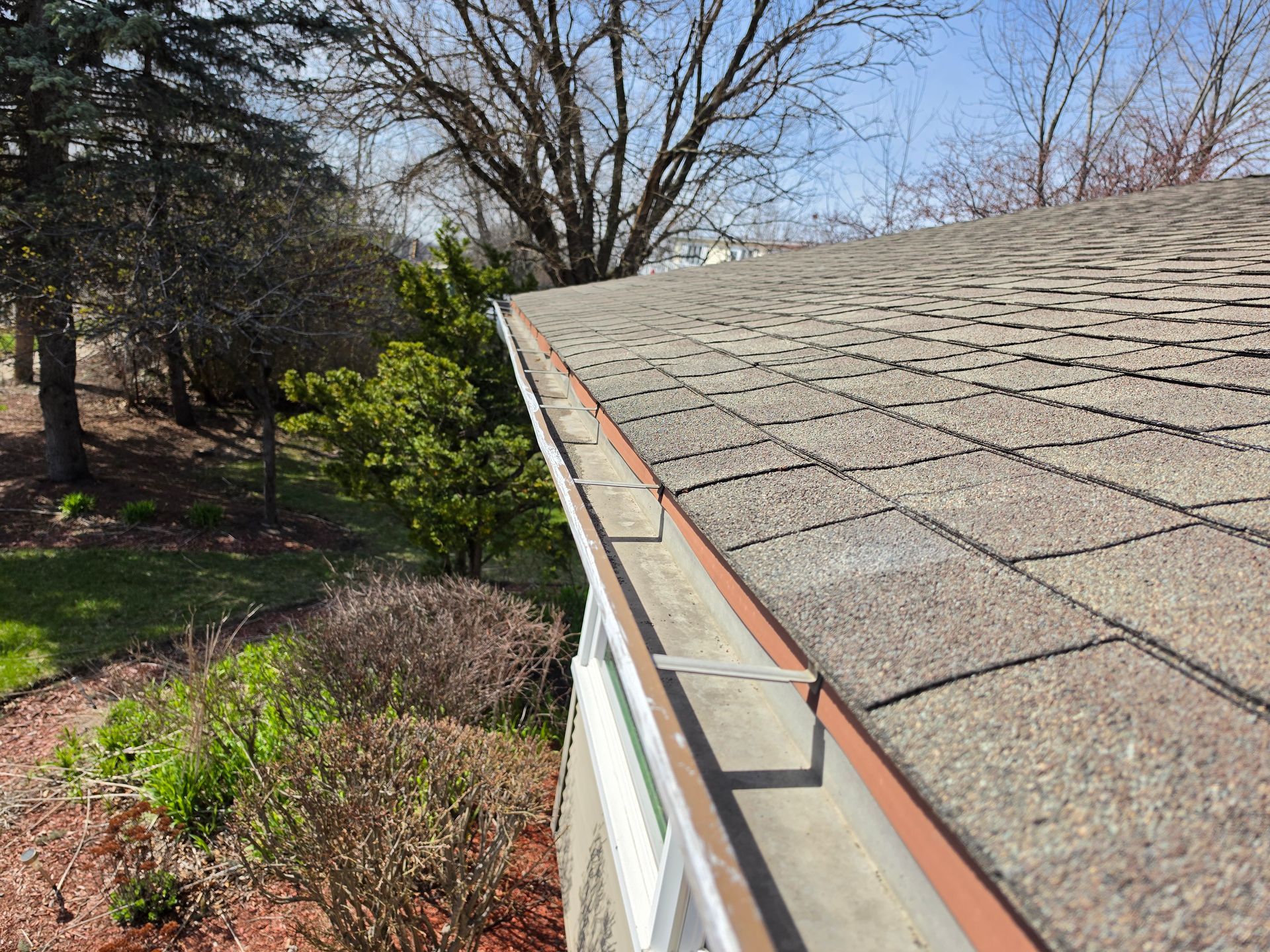 A close-up view of a metal roof gutter attached to the edge of a house roof, with trees and bushes visible in the yard.