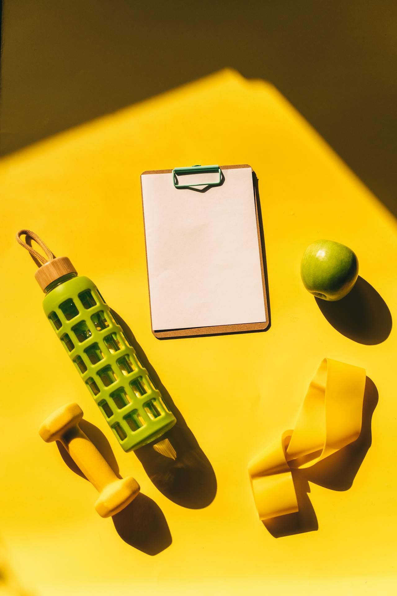 A yellow table with a notebook , dumbbells , a rubber band , an apple and a water bottle.