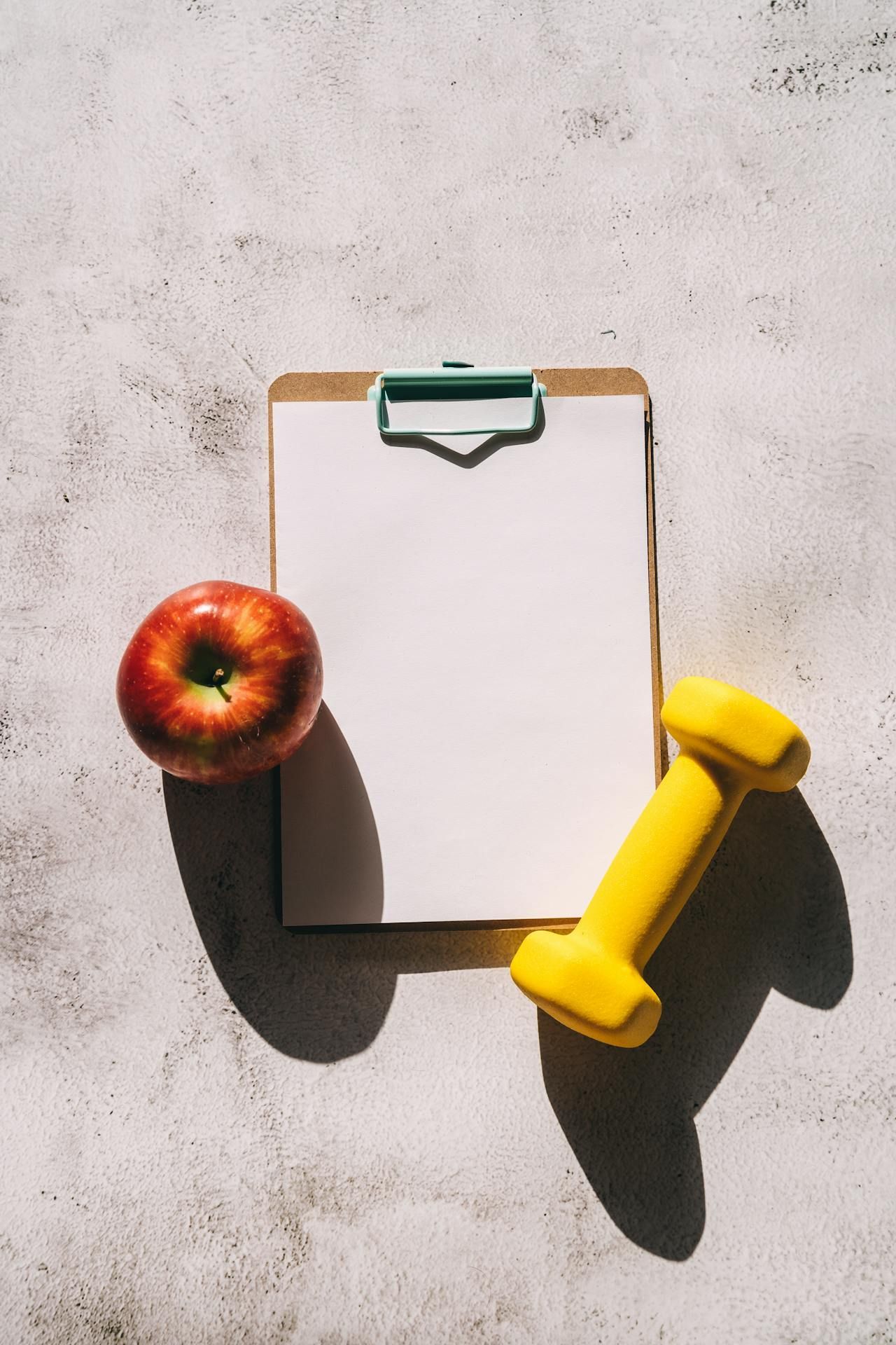 Clipboard, dumbbell, and apple on a table, representing nutrition planning for weight loss.