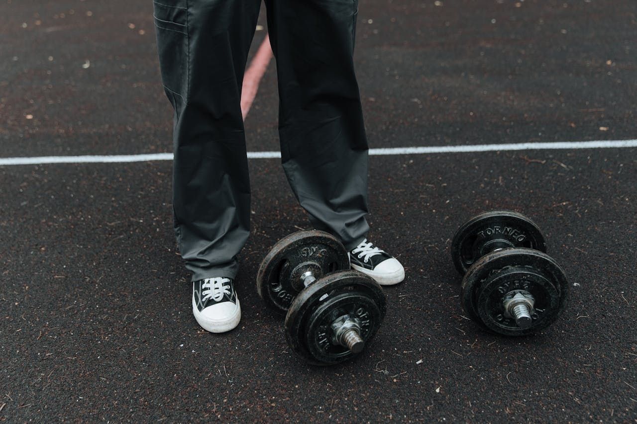 Person in black pants and white sneakers standing near two sets of dumbbells on a dark surface with white lines.