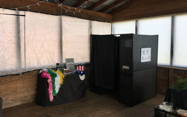 Small indoor booth with black backdrop, rainbow-feathered display table, and a black photo kiosk in a tented room.