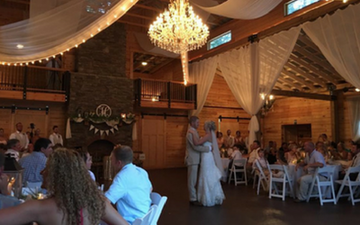 Guests seated at round tables in a warmly lit rustic wedding reception hall with a chandelier and draped ceiling.