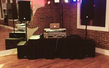 DJ booth setup on a small stage with black tablecloth, speakers, laptop, and colored lights in a brick-walled room