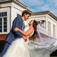 Bride and groom kissing in front of a white building, her veil blowing in the wind