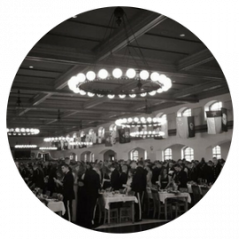 Black-and-white banquet hall with many seated guests and glowing circular chandeliers overhead