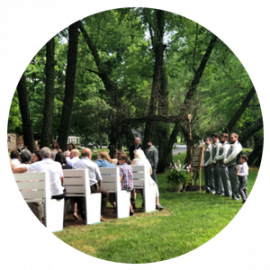 Outdoor wedding ceremony in a wooded park, with guests seated on white chairs and attendants standing nearby