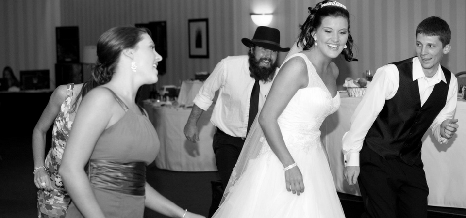 Wedding guests dancing and laughing at a reception, with the bride in a white dress center stage.
