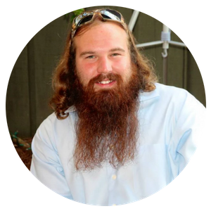 Smiling bearded man in a light blue shirt with sunglasses on his head, seated indoors.