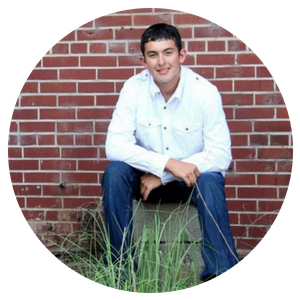 Young man in a white shirt sitting on a concrete block in front of a brick wall and grass.