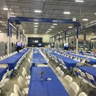 Large indoor event hall with long rows of blue-covered tables and white folding chairs
