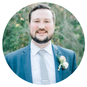 Man in a blue suit and tie smiling outdoors, with a boutonniere on his lapel