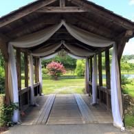 Covered wooden walkway with draped white fabric, opening to a green garden and pink flowering tree.