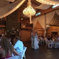 Indoor wedding reception in a rustic hall with a chandelier, white drapes, and guests seated at tables.