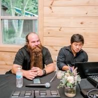 Two men seated at a table with microphones, a laptop, and water bottle in a wooden room