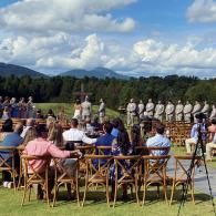 Outdoor ceremony with seated guests facing a line of uniformed people, mountains and clouds in the background