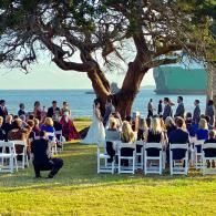 Outdoor wedding ceremony by the water under a large tree, with guests seated on white chairs at sunset