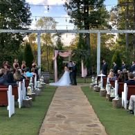Outdoor wedding ceremony with bride and groom under a floral arch, seated guests lining the aisle.