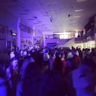 Crowded indoor event space lit in purple and blue, with people standing and walking toward the back